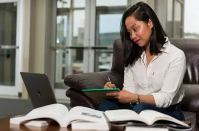 Student studies in a campus common area, taking notes while referencing a laptop and open textbooks. The learning environment supports independent study, research, and academic success.