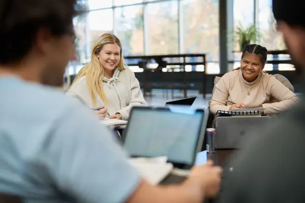 Students sit around a table during a group discussion or study session. Two students smile while listening and taking notes, while laptops and notebooks are used throughout the collaborative learning environment.