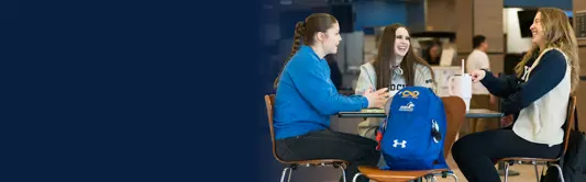 Three students sit around a table in a campus café, chatting and laughing. One holds a mug while a blue backpack rests on a chair in the foreground.
