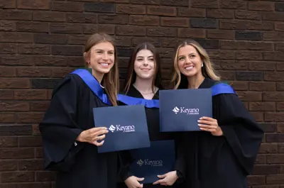 Three Keyano College graduates pose together in graduation gowns, holding diploma folders and smiling in front of a brick wall. The graduates celebrate their academic achievement during a convocation photo session.