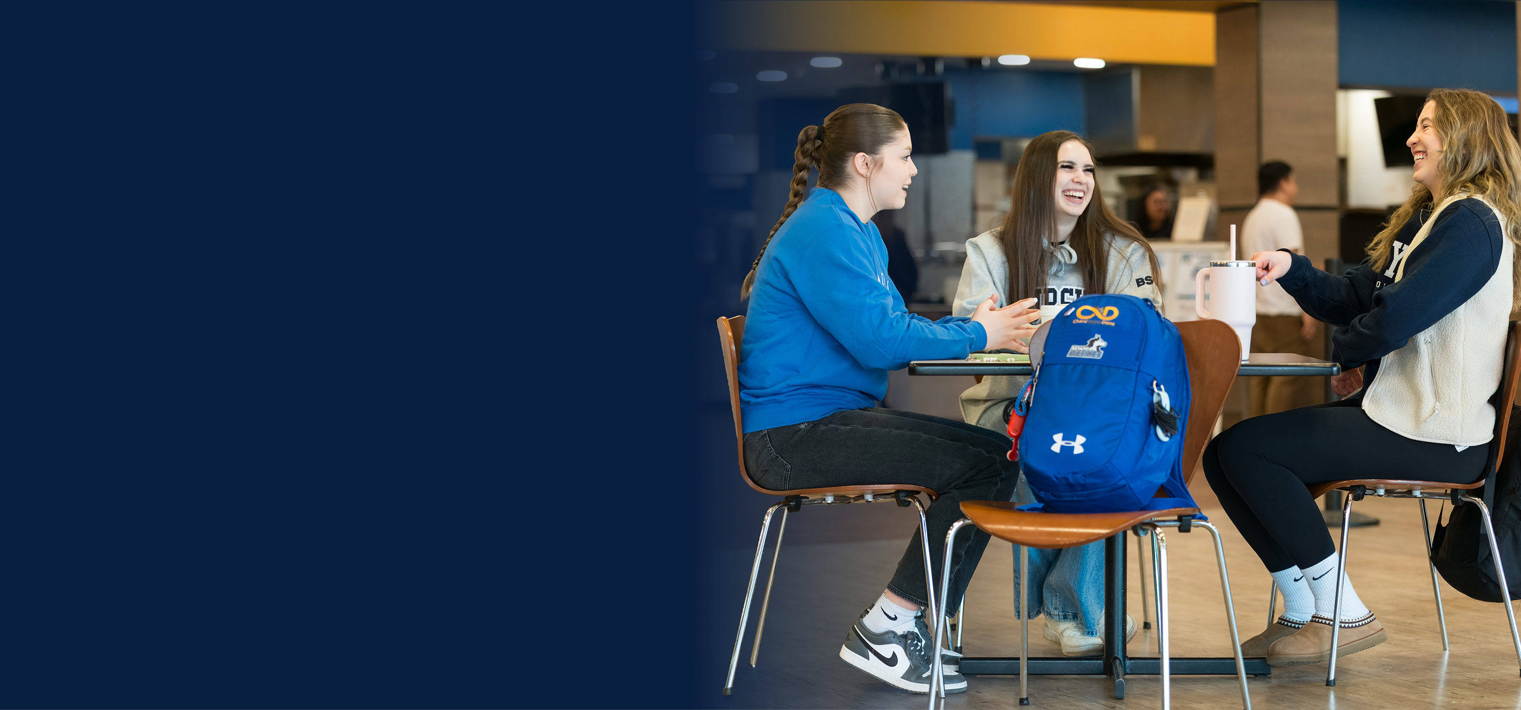 Three students sit around a table in a campus café, chatting and laughing. One holds a mug while a blue backpack rests on a chair in the foreground.